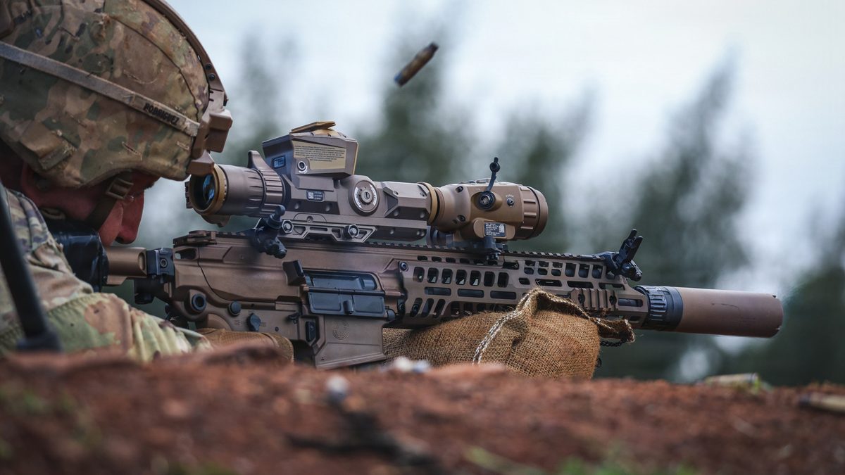 A 25th Infantry Division soldier (helmet name plate &lsquo;Rodriguez&rsquo;) firing the M7 Next Generation Squad Weapon prone from a sandbag rest. The Vortex M157 NGSW-FC optic and SLX suppressor are clearly visible; a brass case is mid-eject above the receiver.