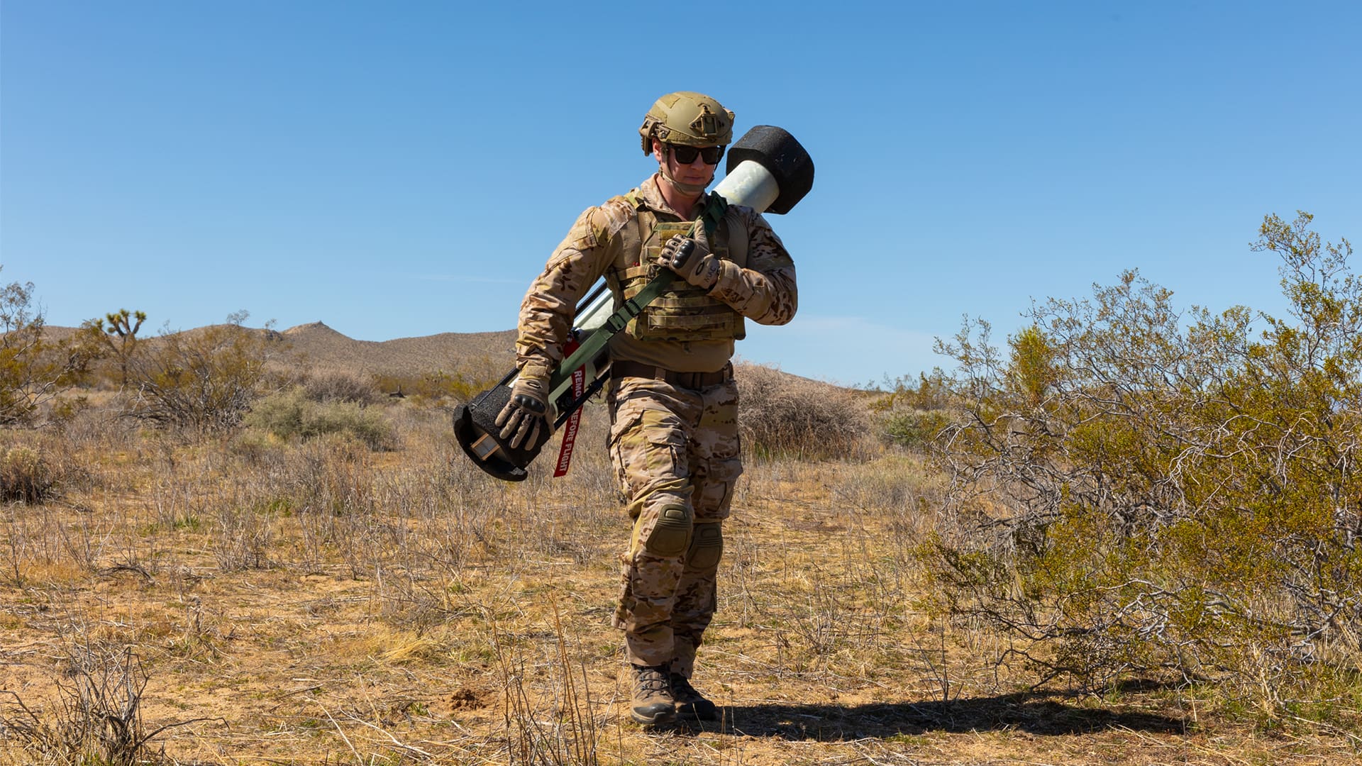 A single soldier carries the Switchblade 400 all-up round (launch tube and munition) across open terrain, demonstrating man-portable employment