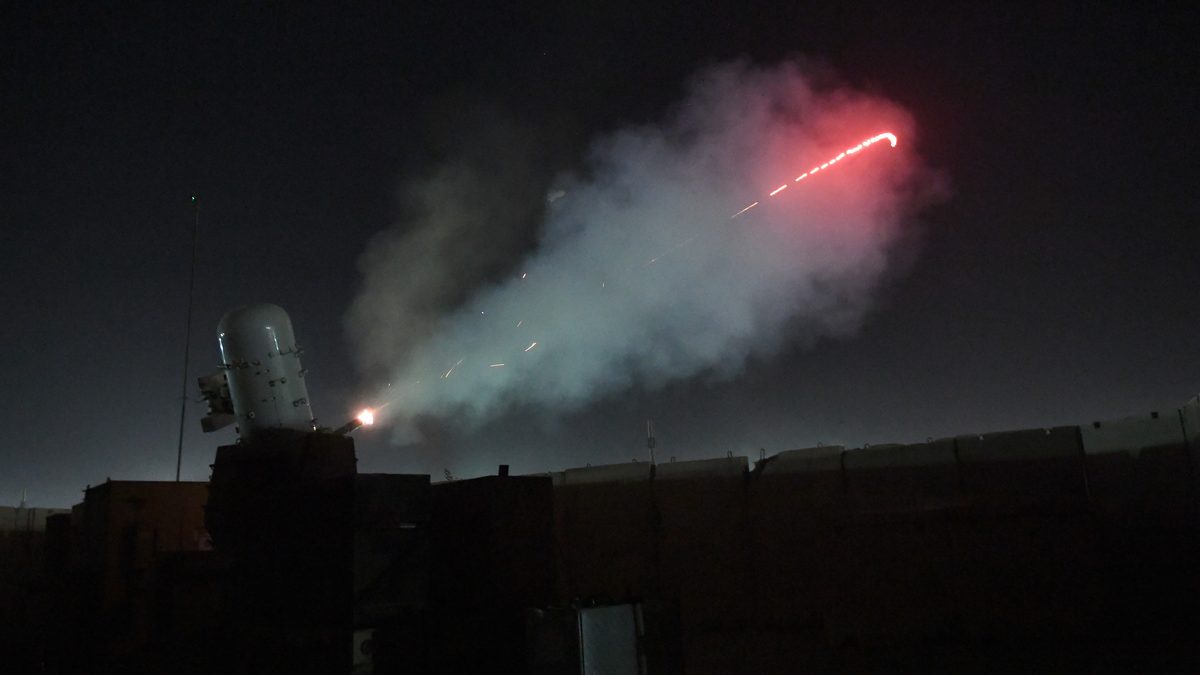 A land-based Phalanx C-RAM gun fires its 20 mm cannon at night, a red tracer arc streaking across the sky.