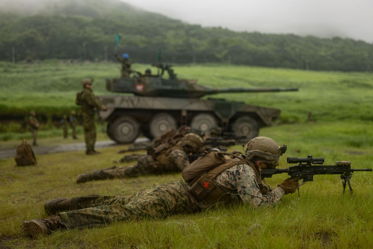 A prone Marine rifleman sights a bipod-mounted M27 during Resolute Dragon 26 in Japan with a Type 16 manoeuvre combat vehicle behind