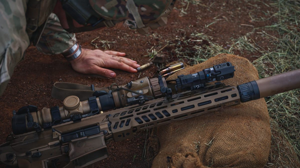 An M7 Next Generation Squad Weapon fitted with a suppressor, rested on a sandbag beside loose 6.8mm cartridges during a live-fire qualification range